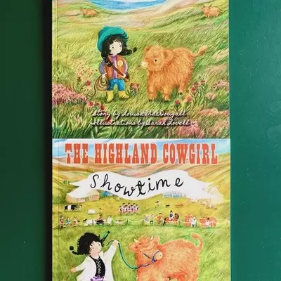 A photo of two picture books , both written by Louisa MacDougall, illustrated by Sarah Lovell and published by Foggie Toddle Books. The Highland Cowgirl and the Highland Cowgirl Showtime. Both books show an illustration of a girl with dark hair in plaits.